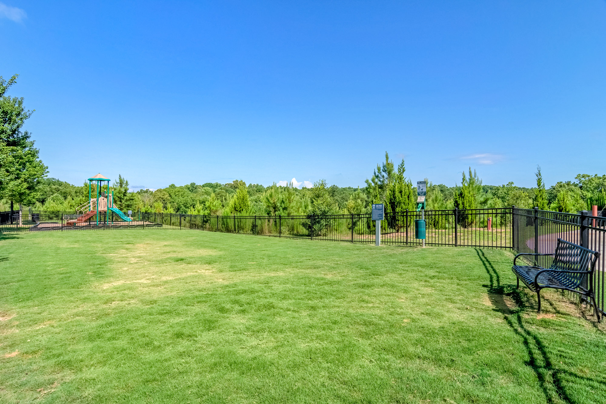 A large fenced in green lawn with a park bench and dog waste system on the right side of the photo.